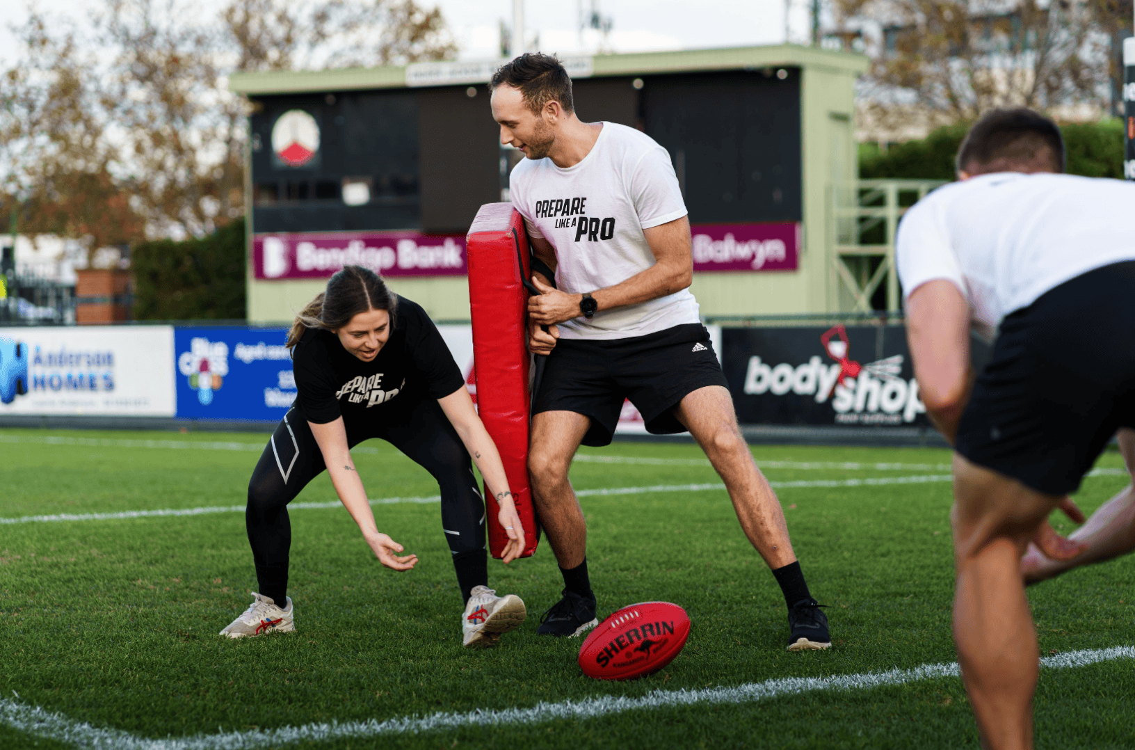 A woman in black sportswear prepares to tackle a pad held by a man in a white shirt during an AFL training session, with a Sherrin football on the ground.