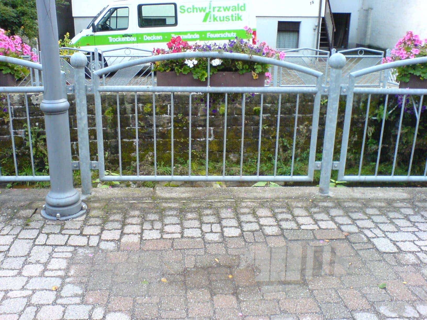 Ein verzinkter Metallzaun von Zaunbau von Stahl- und Metallbau Drost mit dekorativem Bogen, vertikalen Stäben und Kugelaufsätzen, vor einer bemoosten Steinmauer mit Blumenkästen.
