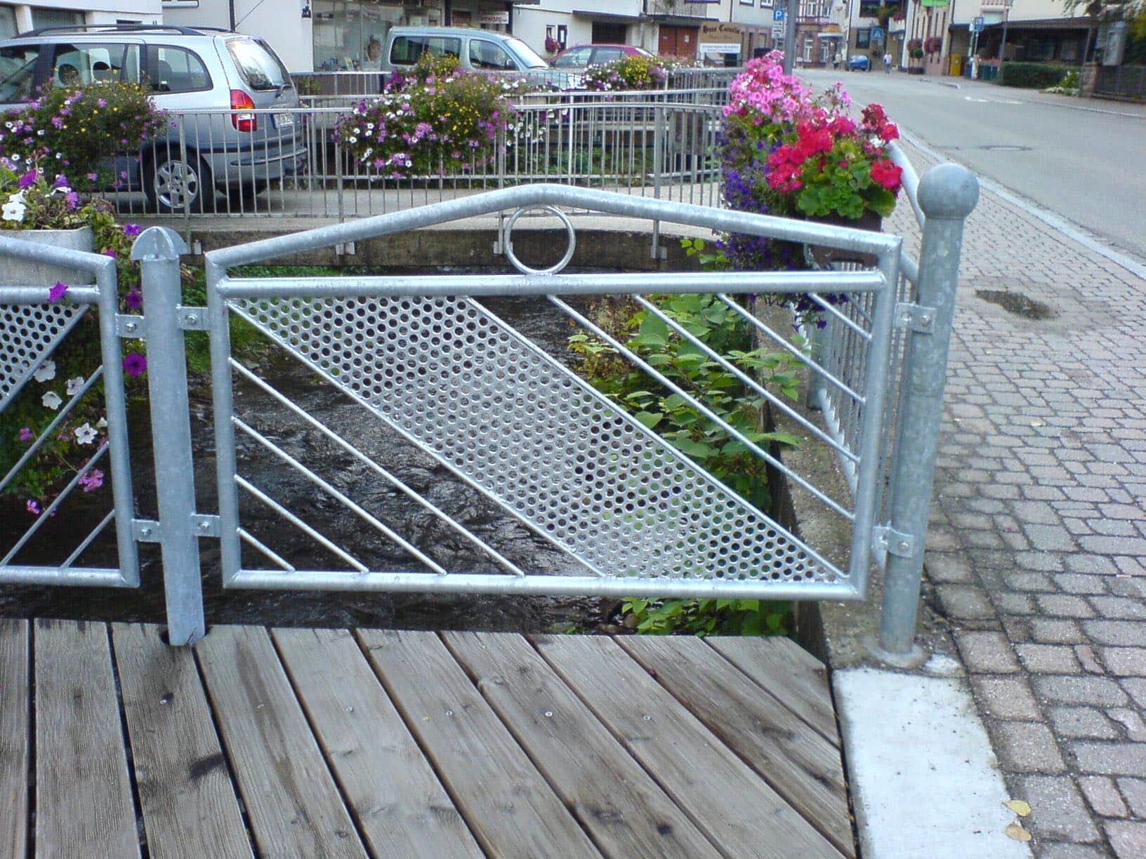 Galvanisiertes Metallgeländer von Zaunbau von Stahl- und Metallbau Drost mit gelochtem Blech und diagonalen Streben, an einem Holzsteg über einem Gewässer, im städtischen Umfeld.