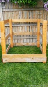 DIY wooden compost bin with wire mesh sides on green grass near a wooden fence and lilac bushes.