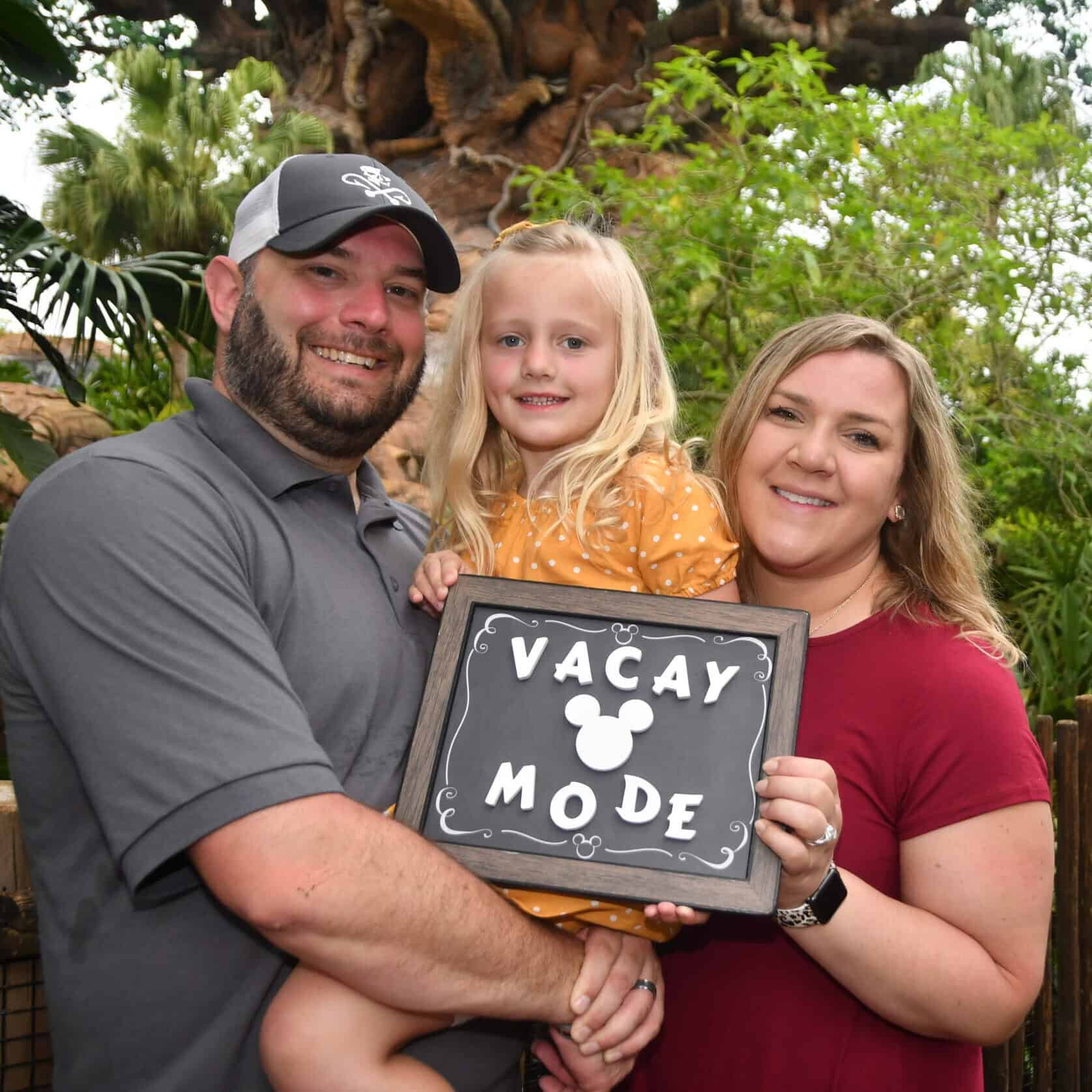 Family enjoying vacation at theme park, holding "Vacay Mode" sign, surrounded by lush greenery and themed decor.