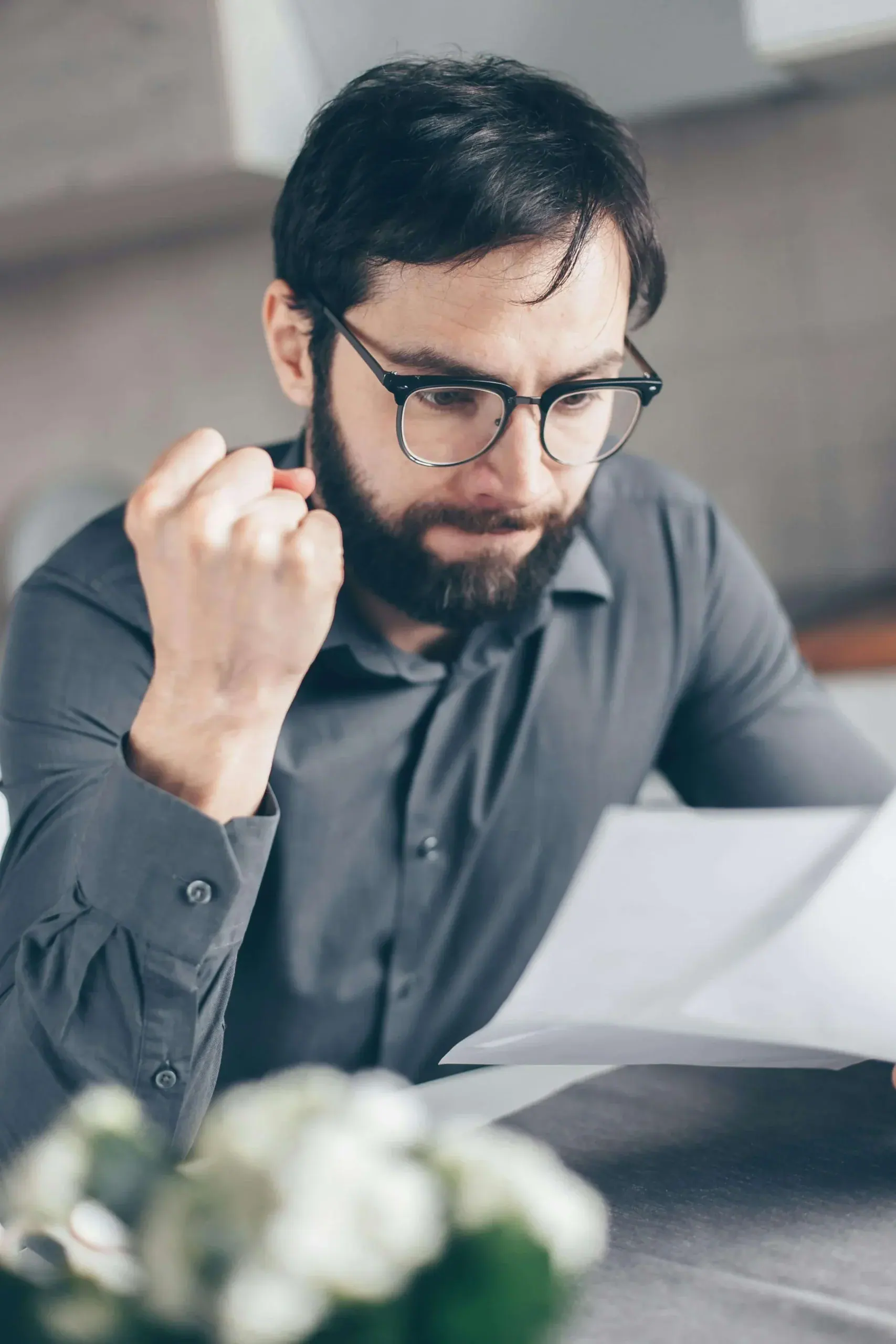 Bearded man with glasses in a gray shirt, looking down at a document he’s holding at a desk, clenching his fist represents emotional connection and support from a therapist for men in Michigan and counselor for men in Detroit, MI.