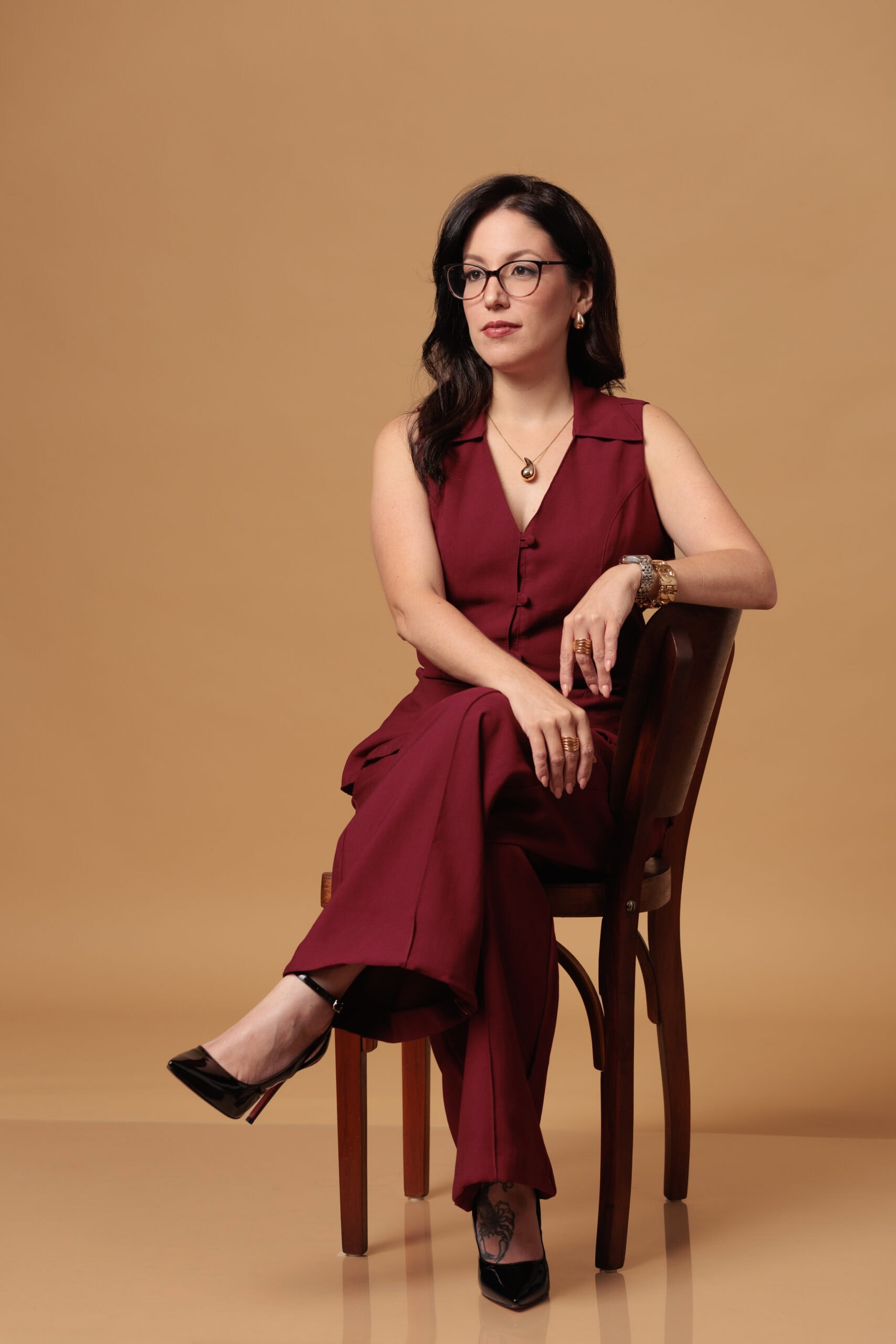 Retrato profissional de uma mulher de óculos e conjunto vinho, sentada numa cadeira de madeira com as pernas cruzadas, em fundo bege, feito pelo Alê no estúdio em BH
