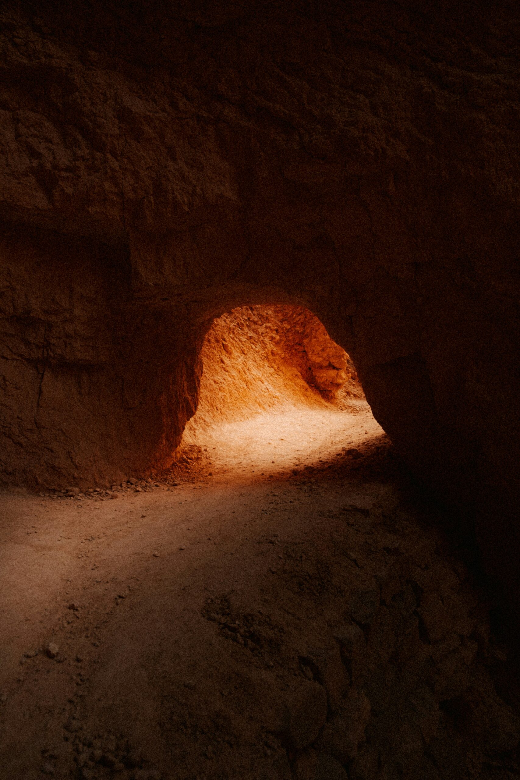 Narrow rocky tunnel with an illuminated dirt path and reddish-brown walls.