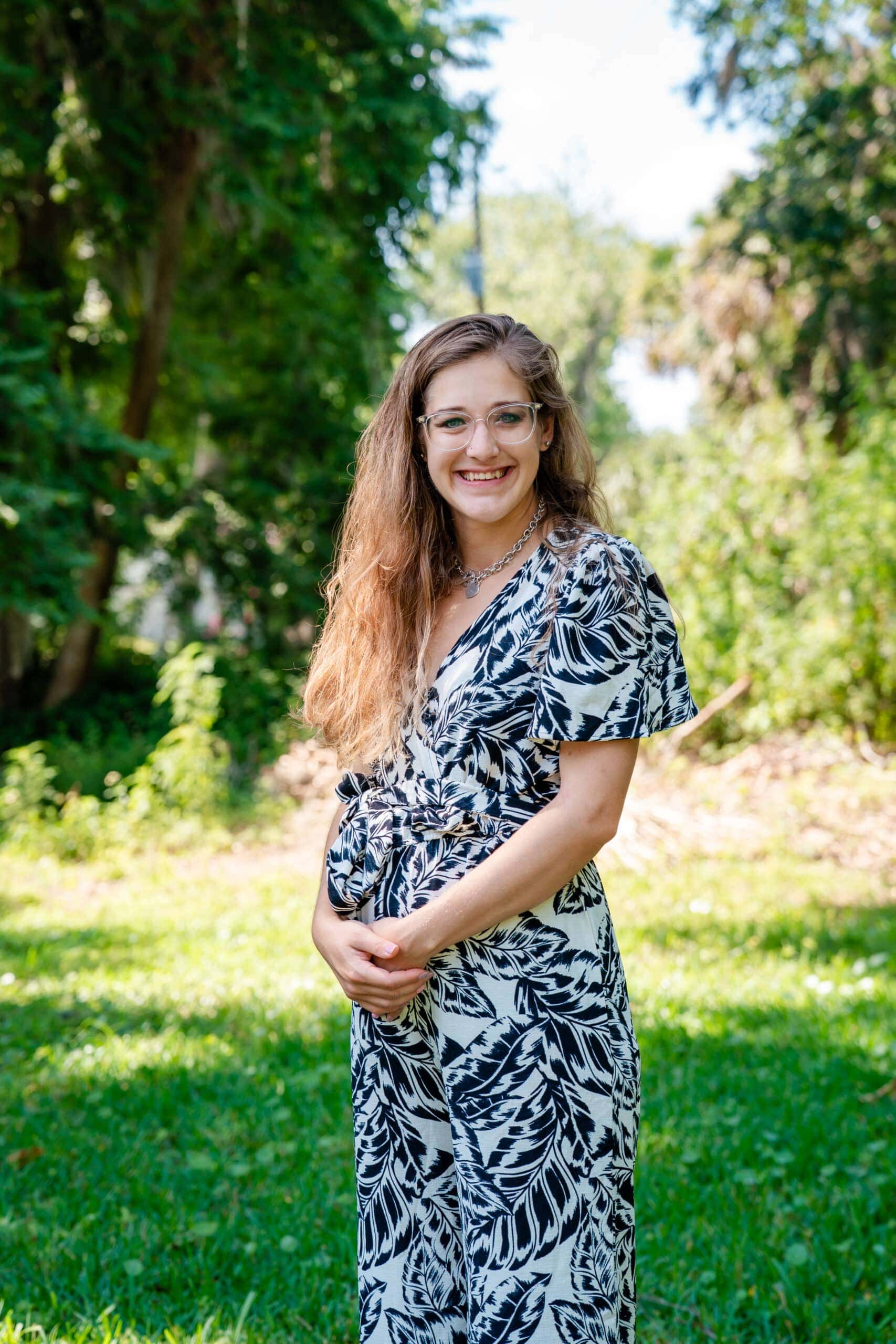 Woman in a black and white dress standing in a grassy outdoor area, smiling and wearing glasses. MPR.
