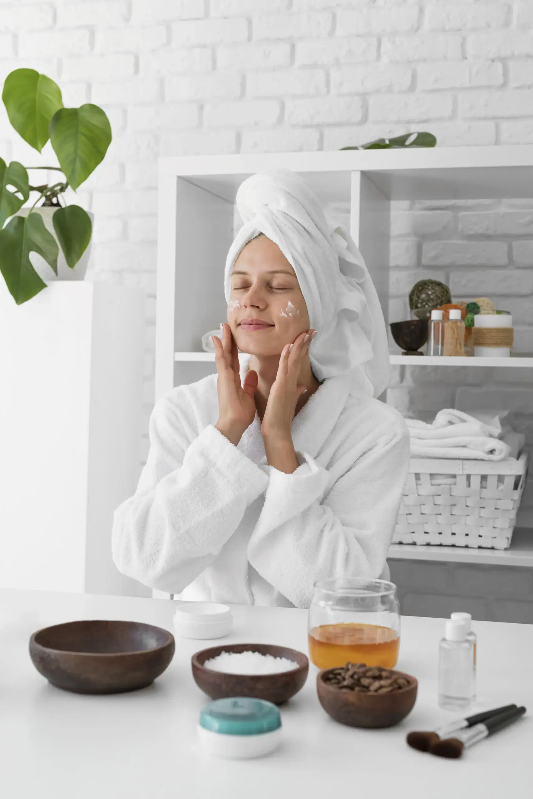 Personal Care- A woman in a white bathrobe and head towel applies face cream while seated at a table featuring natural beauty ingredients. The scene includes wooden bowls of coffee beans and salts, a jar of honey, and skincare tools set against a bright, background.