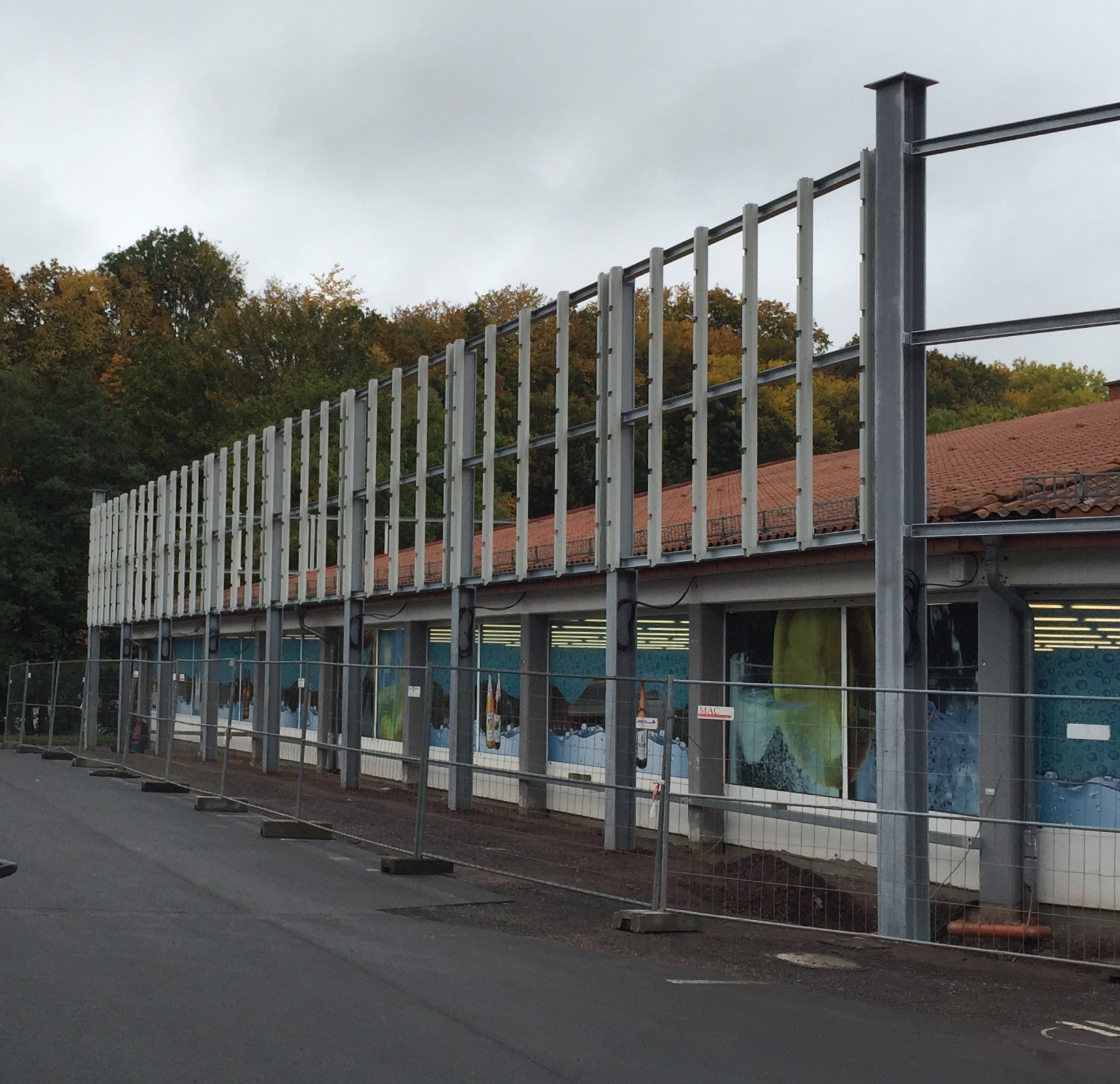 Stahlkonstruktion von Stahl- und Metallbau Drost an der Fassade eines Gebäudes. Das Gerüst aus vertikalen und horizontalen Stahlträgern steht vor einem Gebäude mit rotem Ziegeldach. Bauzäune im Vordergrund.