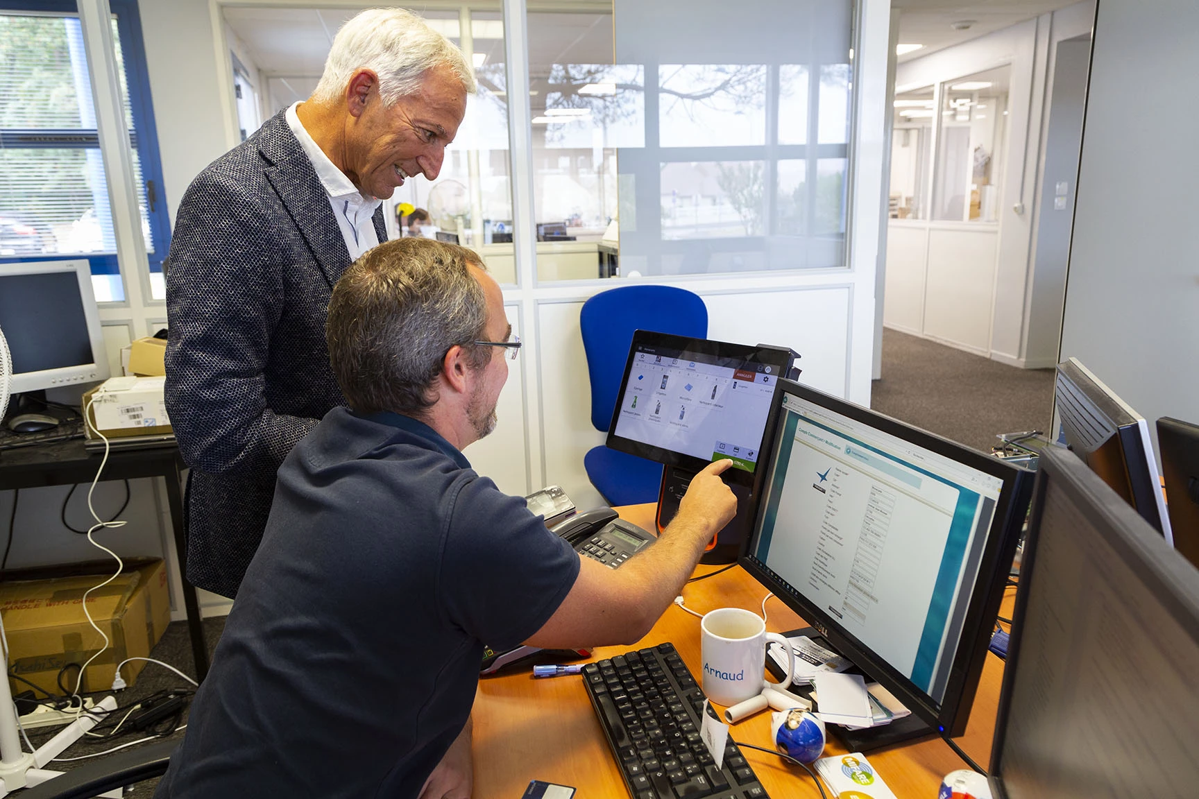 Deux hommes travaillent ensemble dans un bureau. L'un utilise un ordinateur tandis que l'autre observe par-dessus son épaule