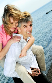 Smiling couple by the water, woman in pink shirt playfully on man’s back, joyful moment representing dentistry.