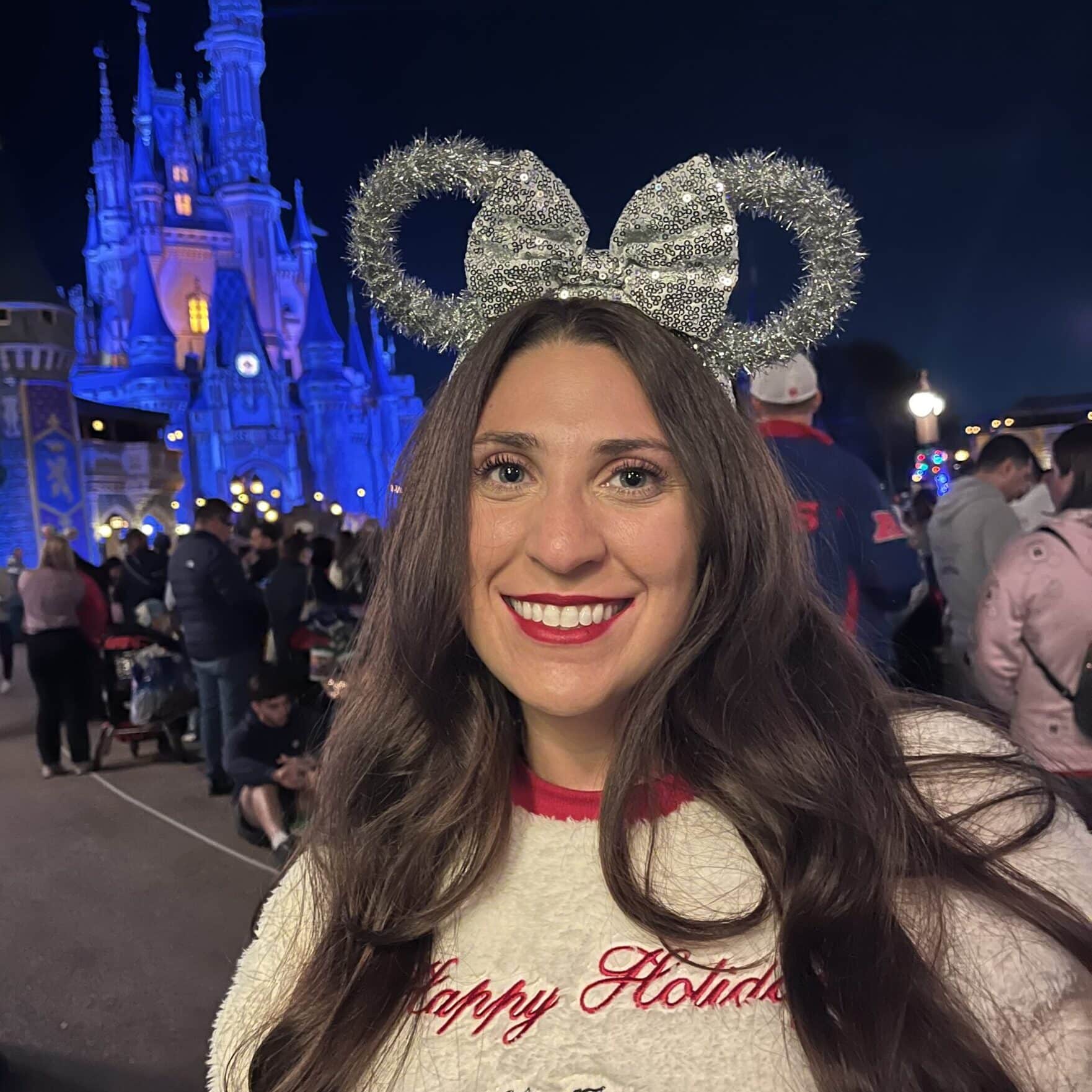 Woman smiling at Disney park with castle in background, wearing holiday sweater and Mickey ears.