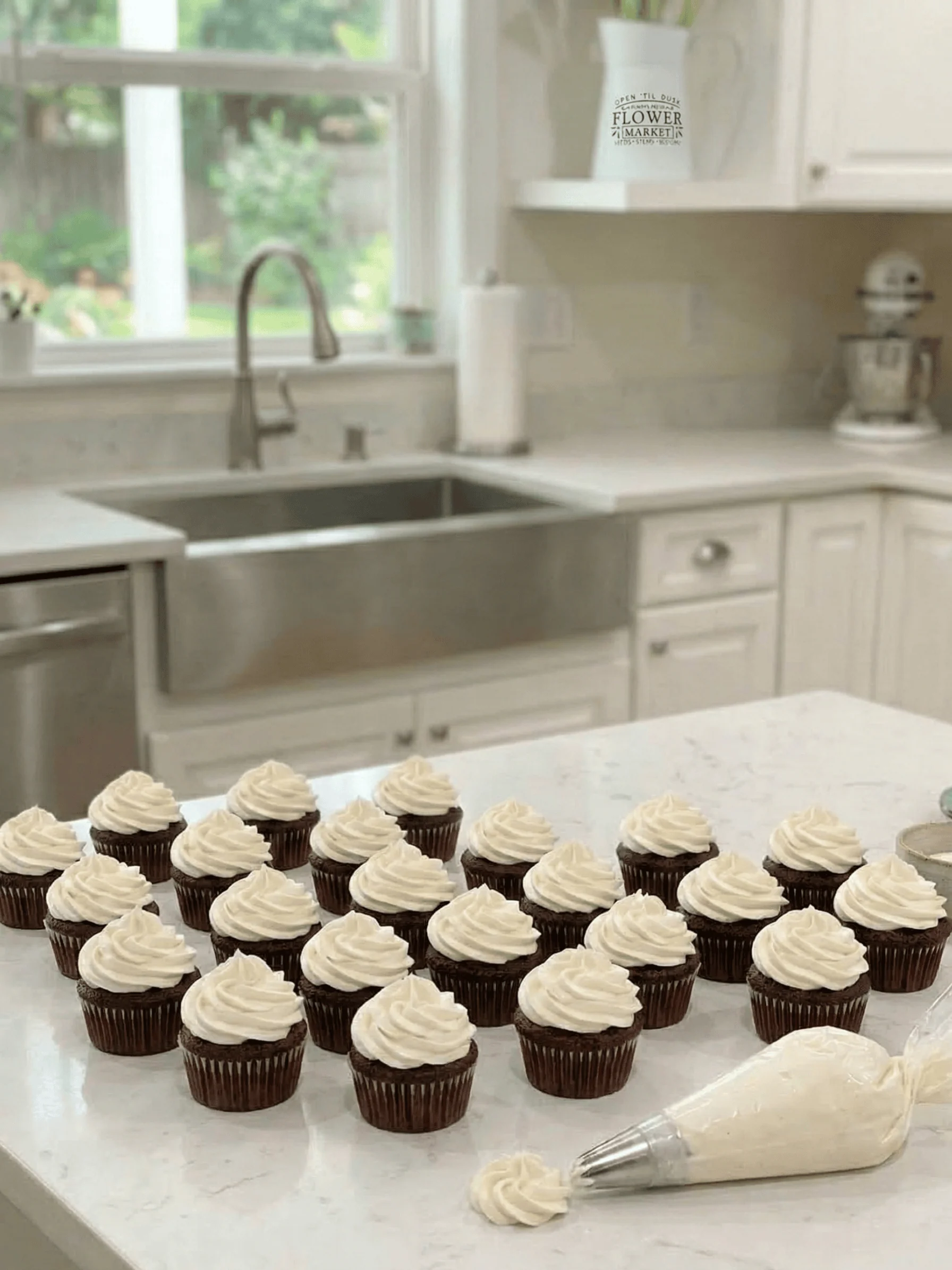 A long view of chocolate cupcakes on a white marble kitchen island with a clear piping bag filled with vanilla frosting in the foreground.
