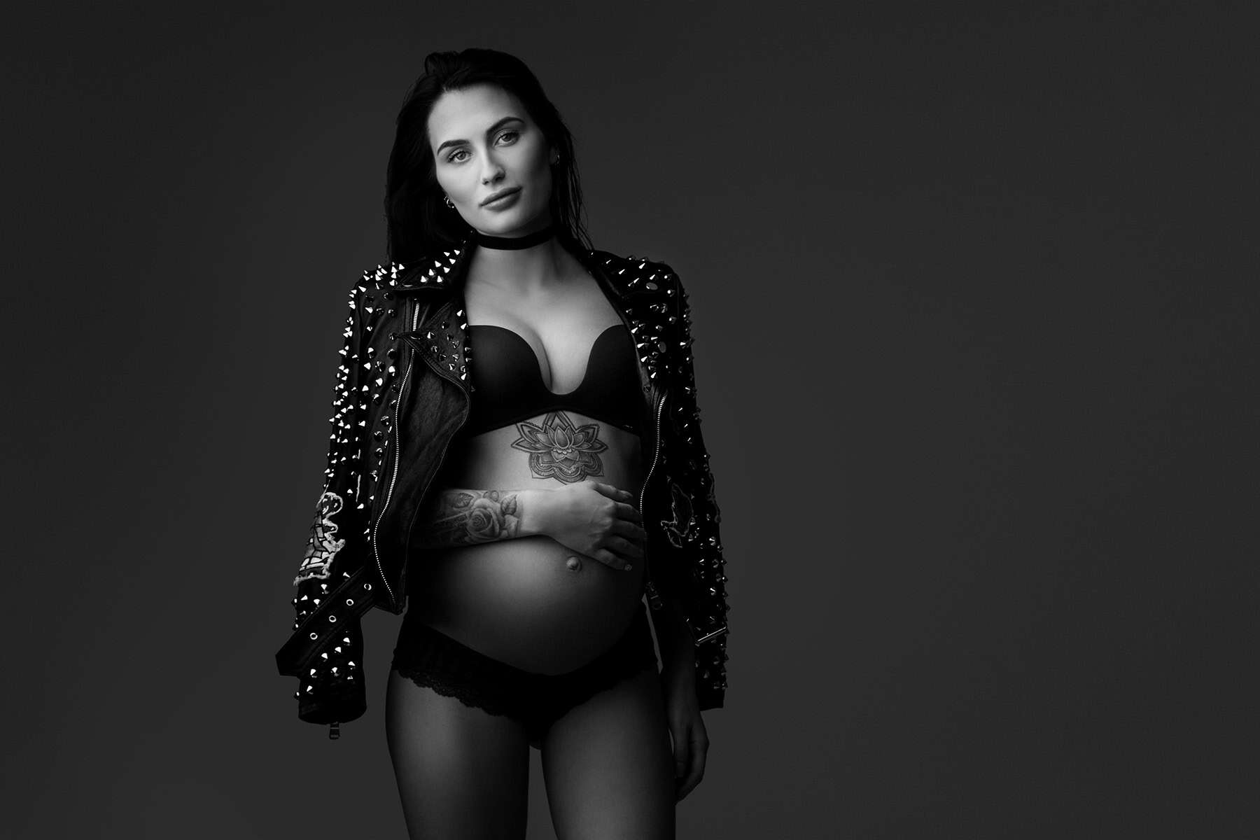 Elegant gravidfoto av en gravid kvinne i sexy antrekk, svart og hvitt, med tatoveringer og skinnjakke med metallspikler, som fremhever hennes livmor og prenatal skjønnhet.