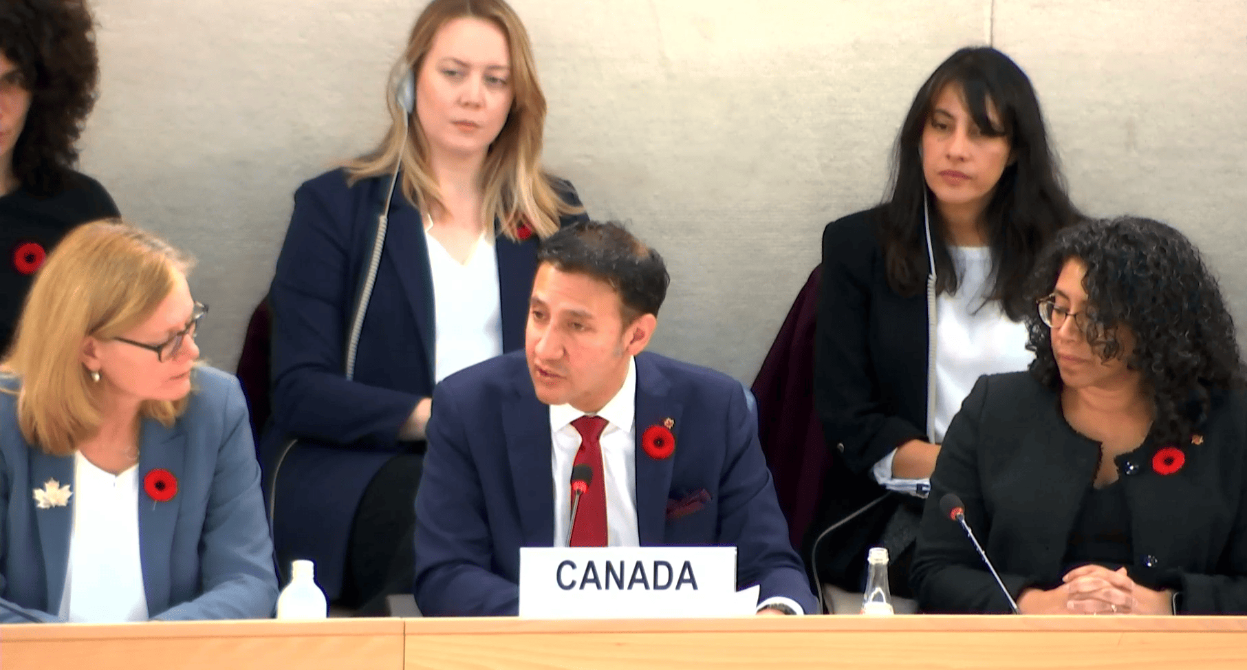 A man wearing a blue suit jacket and a red tie speaks into a microphone while seated while two women beside him look on