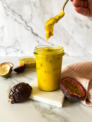 passion fruit curd in a jar on a marble coaster with a spoon held by a hand, around passion fruits, towel and more curd in a bowl behind, on a marble background