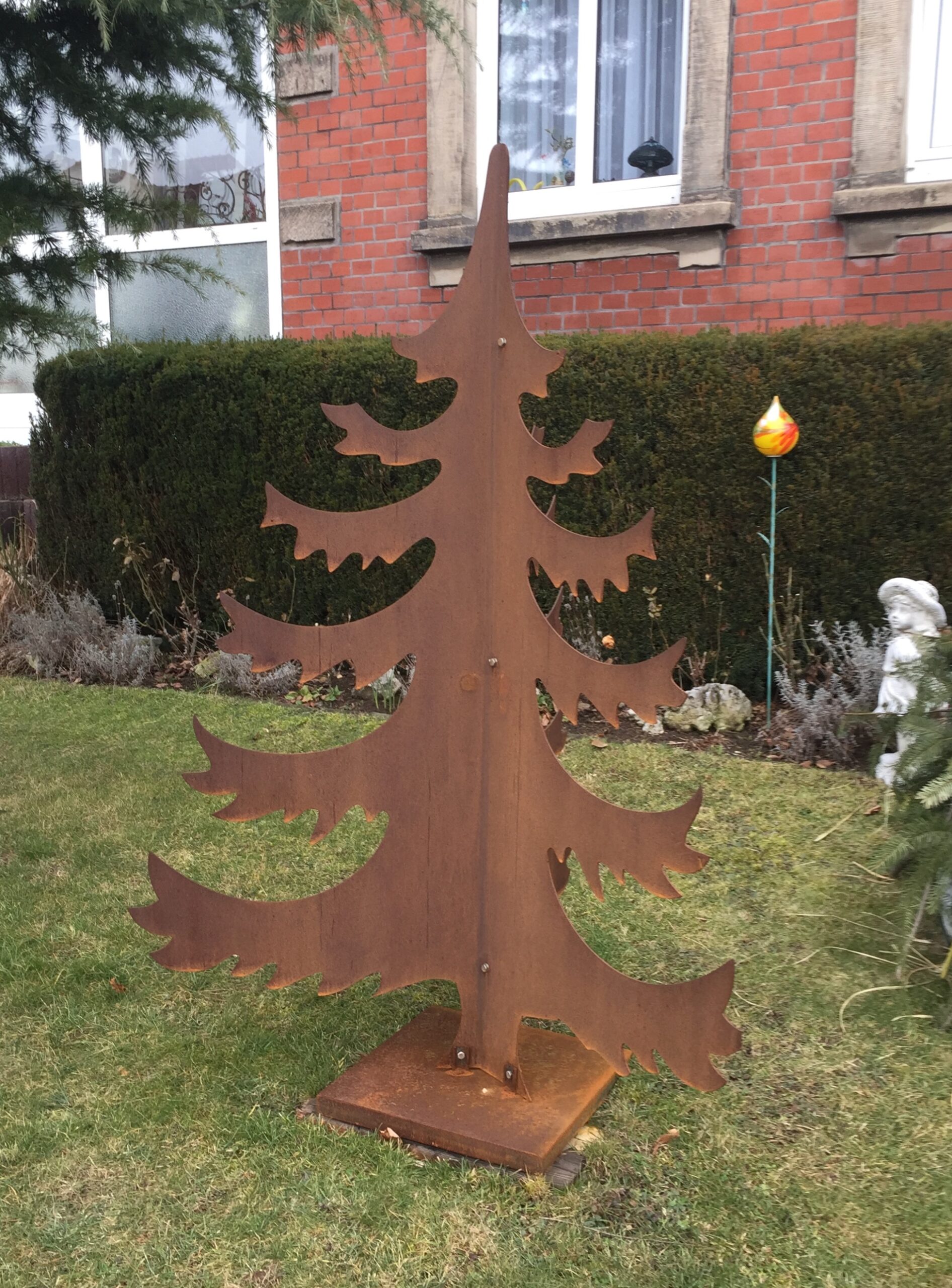 Dekorative Tannenbaum-Skulptur aus Cortenstahl für den Garten, eine Sonderanfertigung von Stahl- und Metallbau Drost. Die Skulptur zeigt einen stilisierten Weihnachtsbaum mit natürlicher Rostpatina, aus zwei ineinandergesteckten Metallplatten geformt und auf einem quadratischen Sockel im Gras platziert.