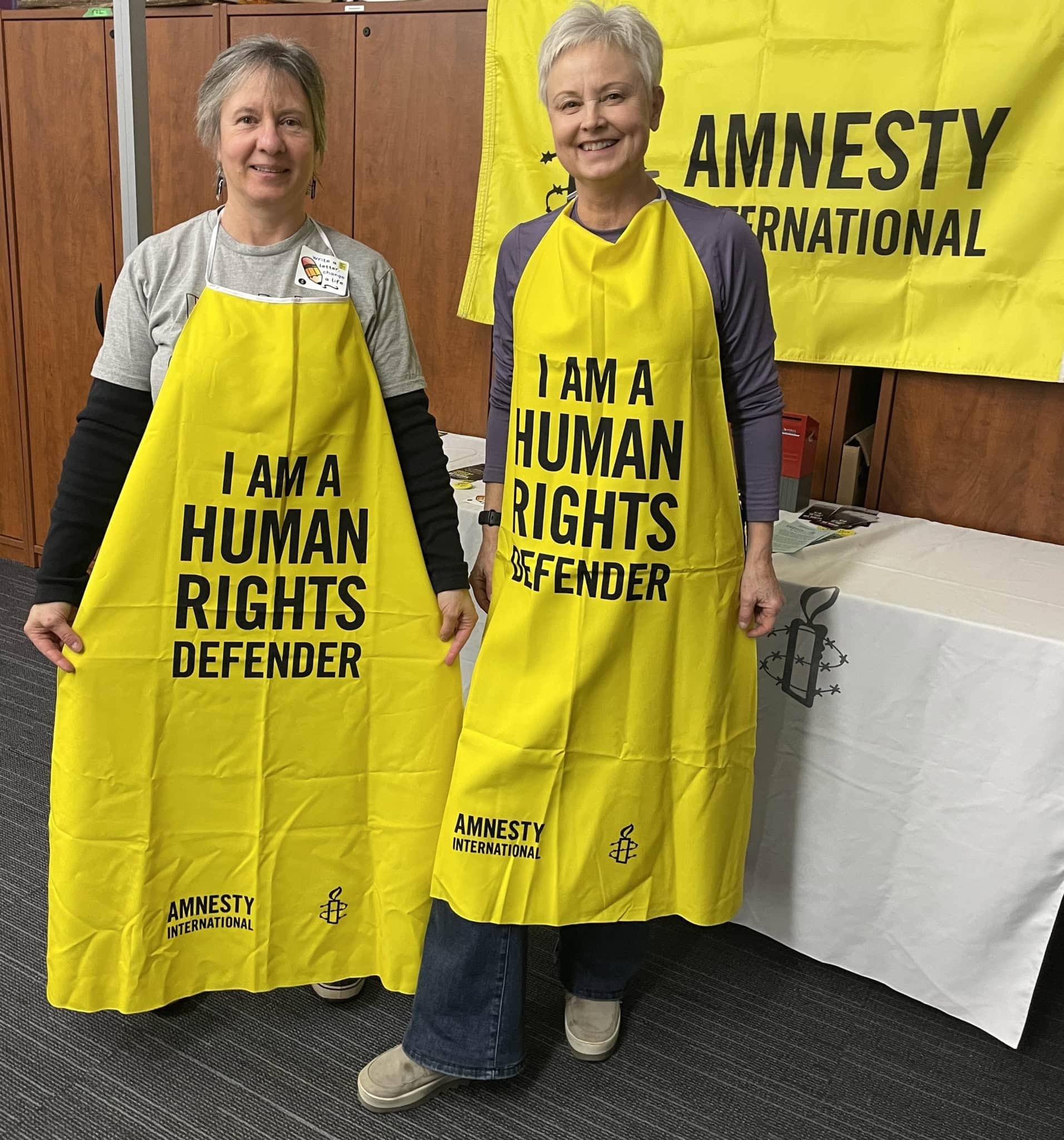 “Two Amnesty International volunteers standing side by side, wearing bright yellow ‘I am a human rights defender’ aprons at a Write for Rights event.”