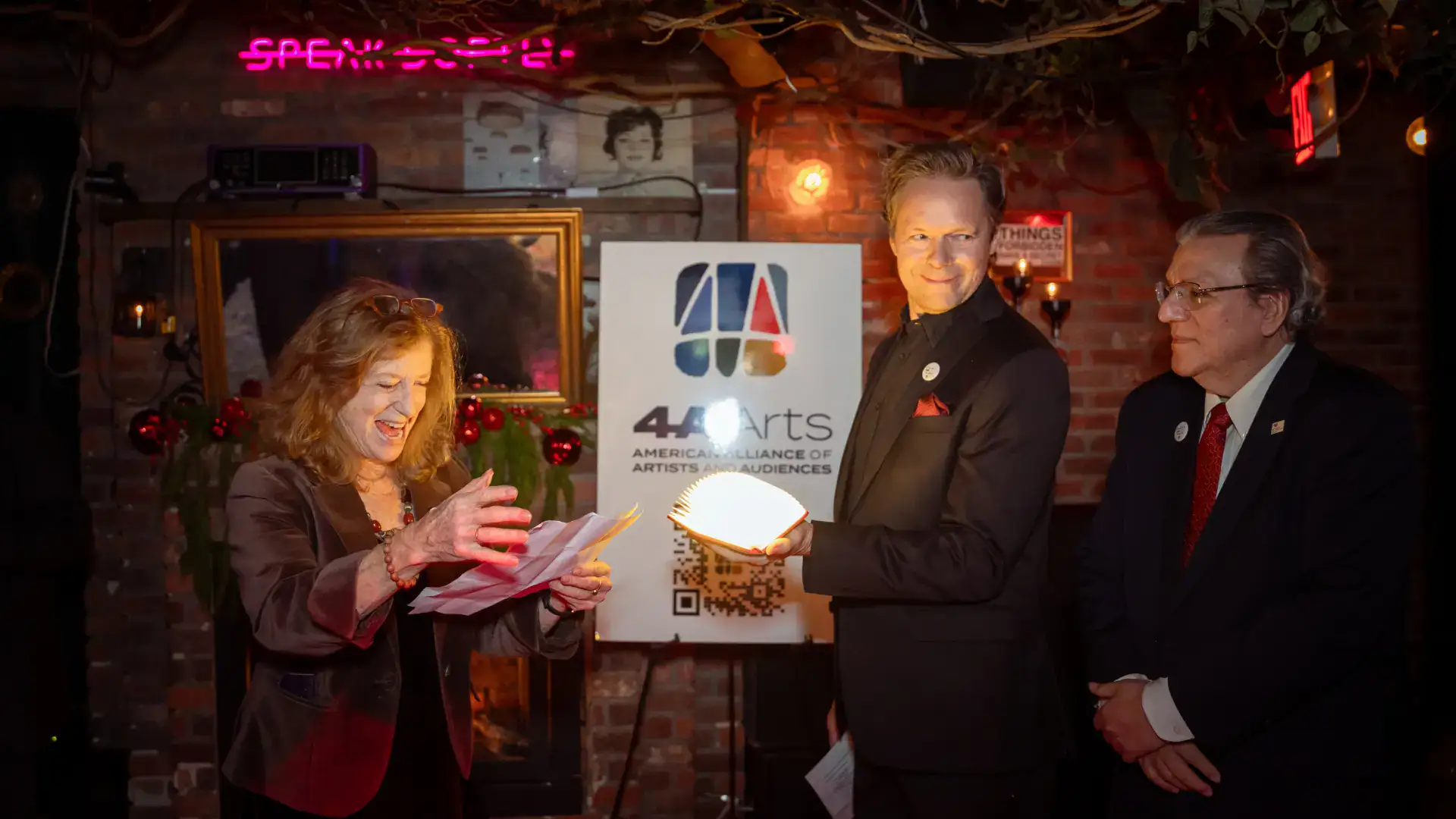 Joy Javits, a woman with strawberry blond hair reads from a paper and laughs while Gavin Lodge and Luis Cancel look on.