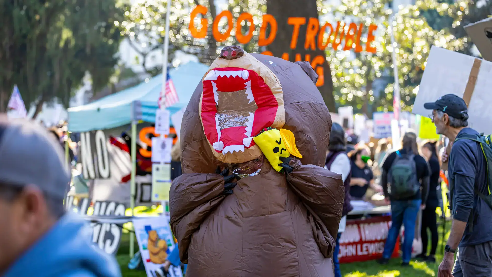 A person in an inflatable bear costume pretends to eat a toy taco. In the back is a tent set up at a protest.