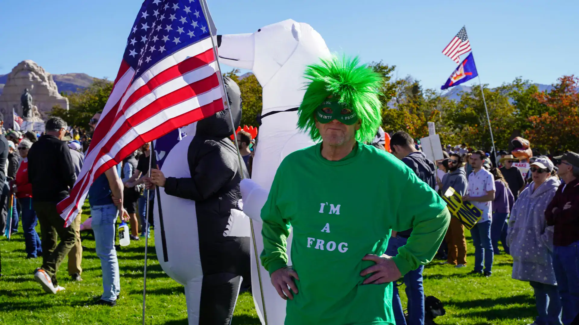 A man in a green wig and a green mask wearing a shirt that says "I'm a frog" stands in front of an American flag and another protestor in an inflatable polar bear costume