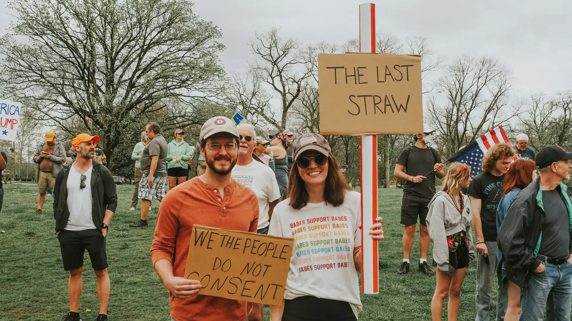 A woman at a protest holds a giant straw and a sign saying "The Last Straw." A man stands next to her with a sign saying "We the people do not consent."
