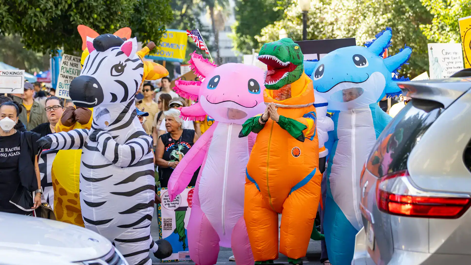 Several protestors are in silly inflatable costumes, including a zebra, dinosaur, and two axolotls