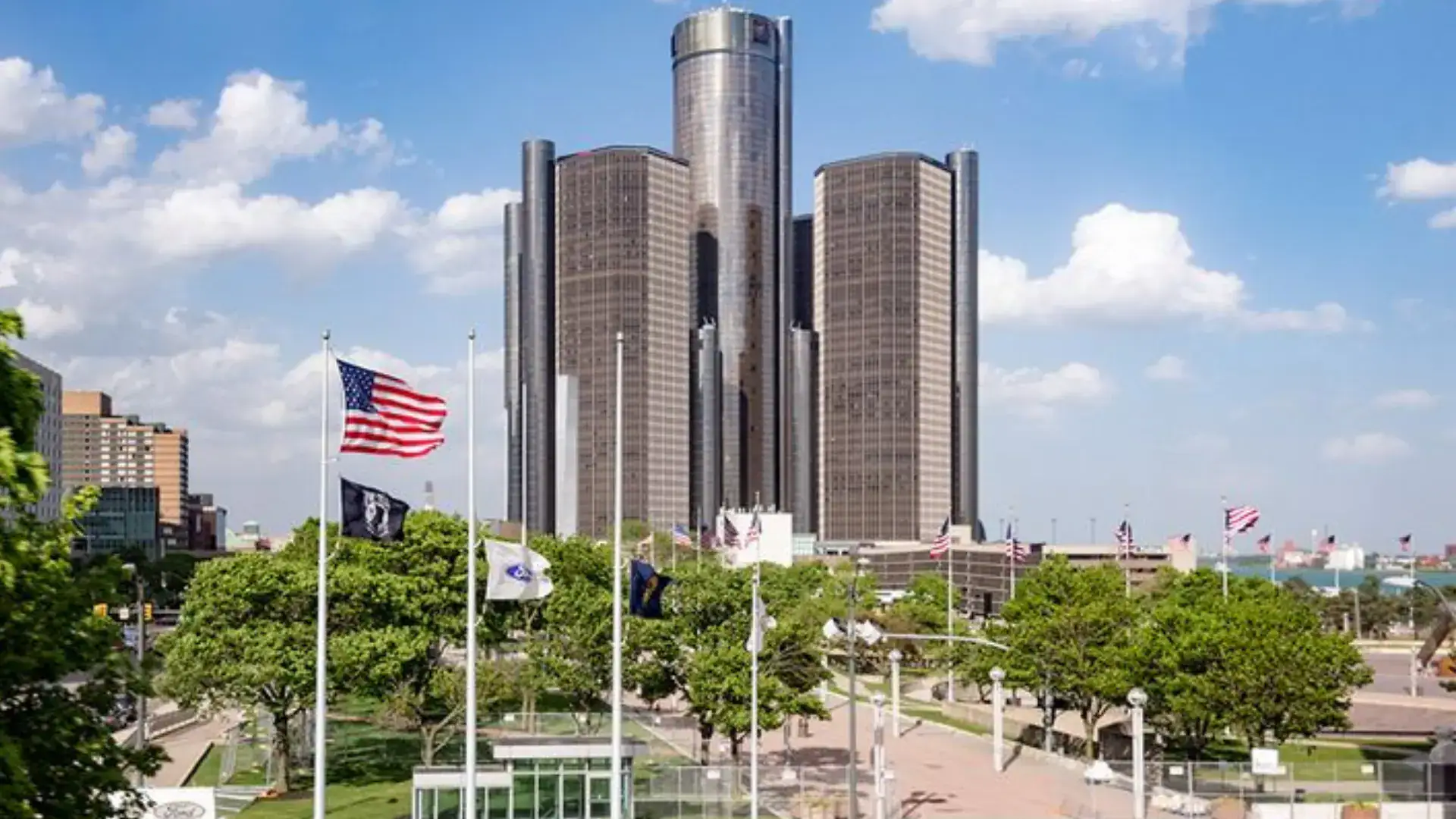 Renaissance Center towers in downtown Detroit with flags, tree-lined plaza, and clear sky highlighting the city’s skyline.