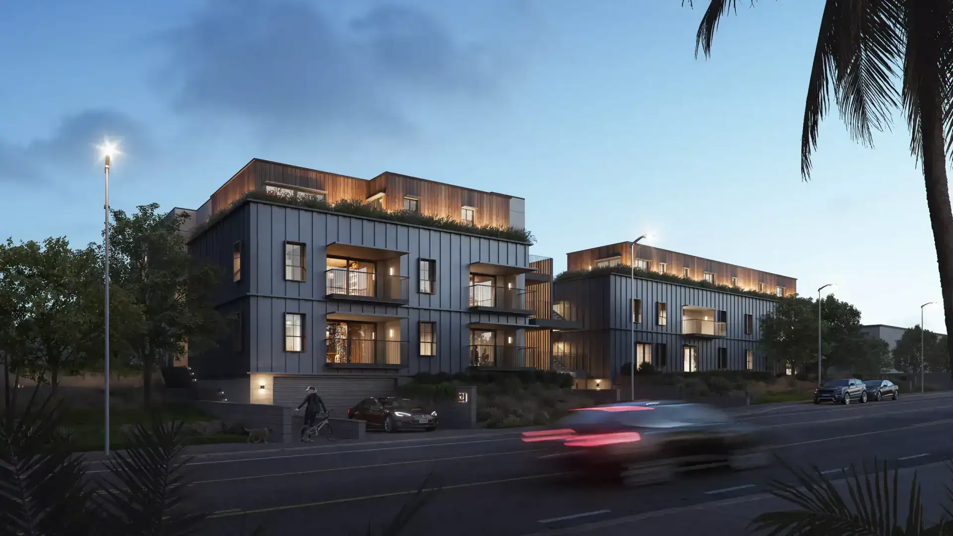 Modern multi-story residential building with mixed materials, featuring metal panel facades, wooden accents, and outdoor terraces illuminated at dusk.