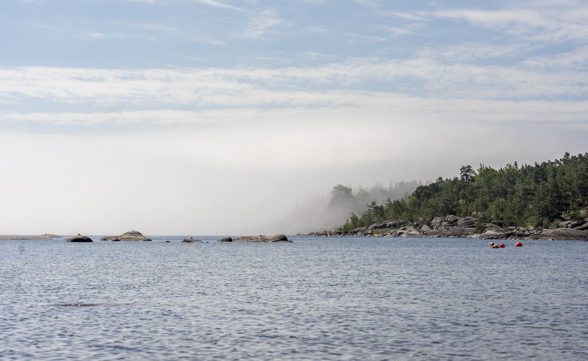 Vy över skärgård med klippor, skog och hav, där dimma rullar in över horisonten under en klarblå himmel.