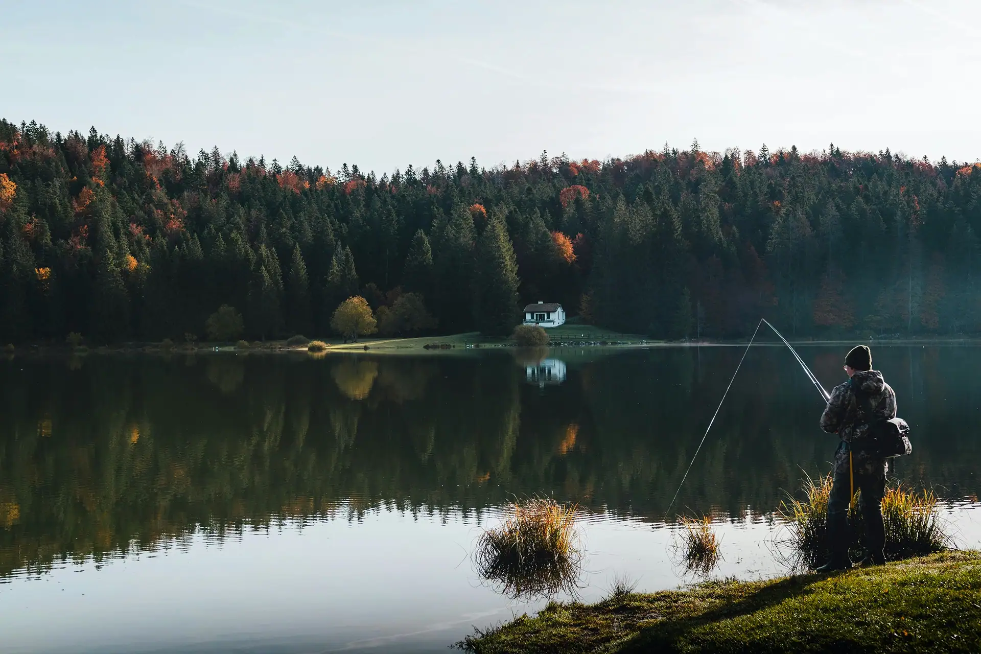 An angler on the shore of a glassy lake reflecting a colorful autumn forest and a lone white house.