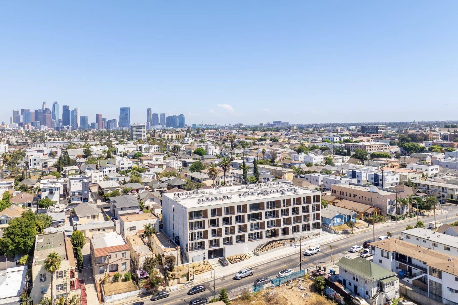 Modern multi-story apartment building with a minimalist design, large glass balconies, and a flat roof, situated in a densely populated urban neighborhood.