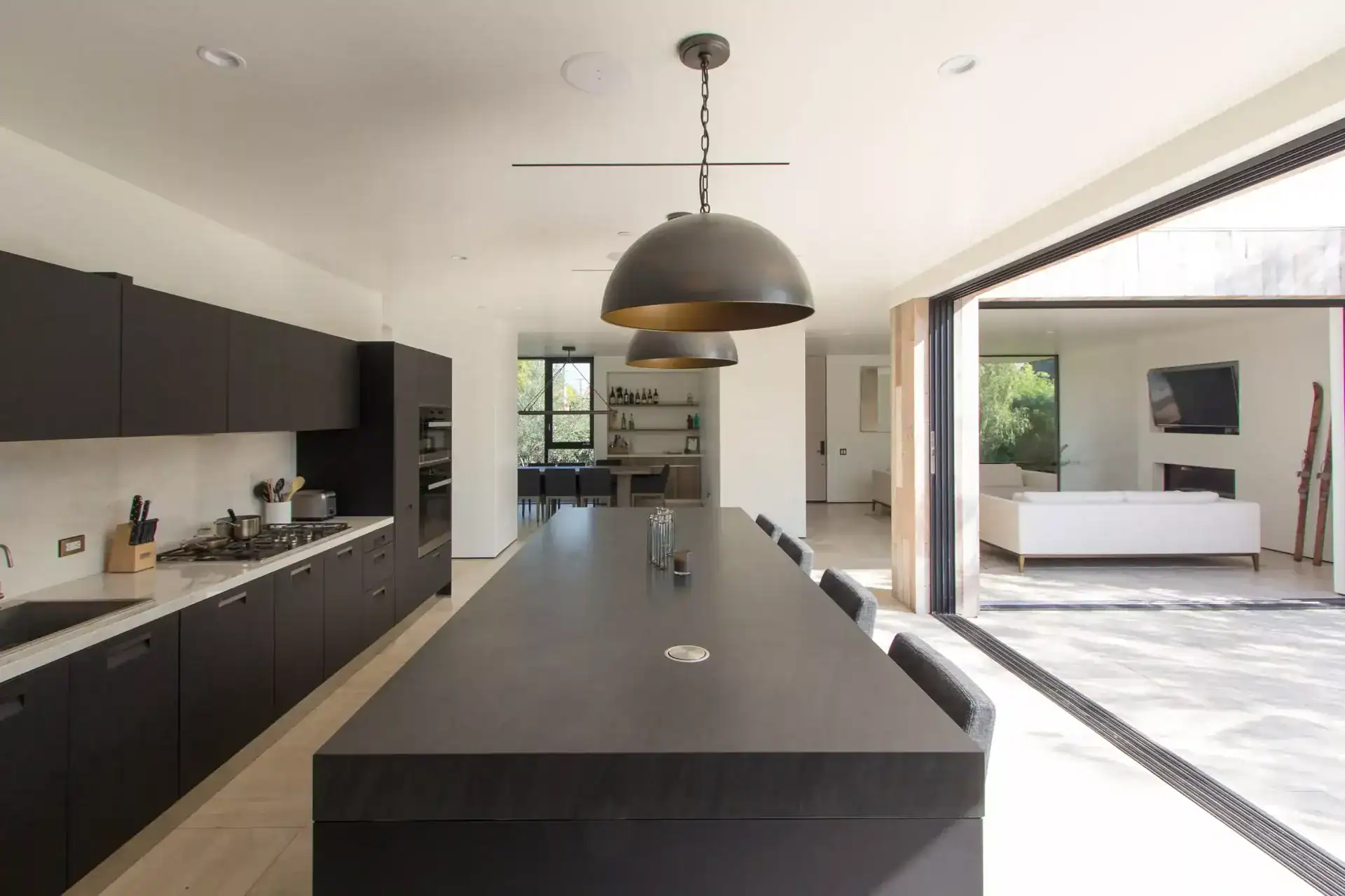 Sleek modern kitchen with dark cabinetry, minimalist island, and pendant lighting, open to a bright living area with large windows and sliding doors.