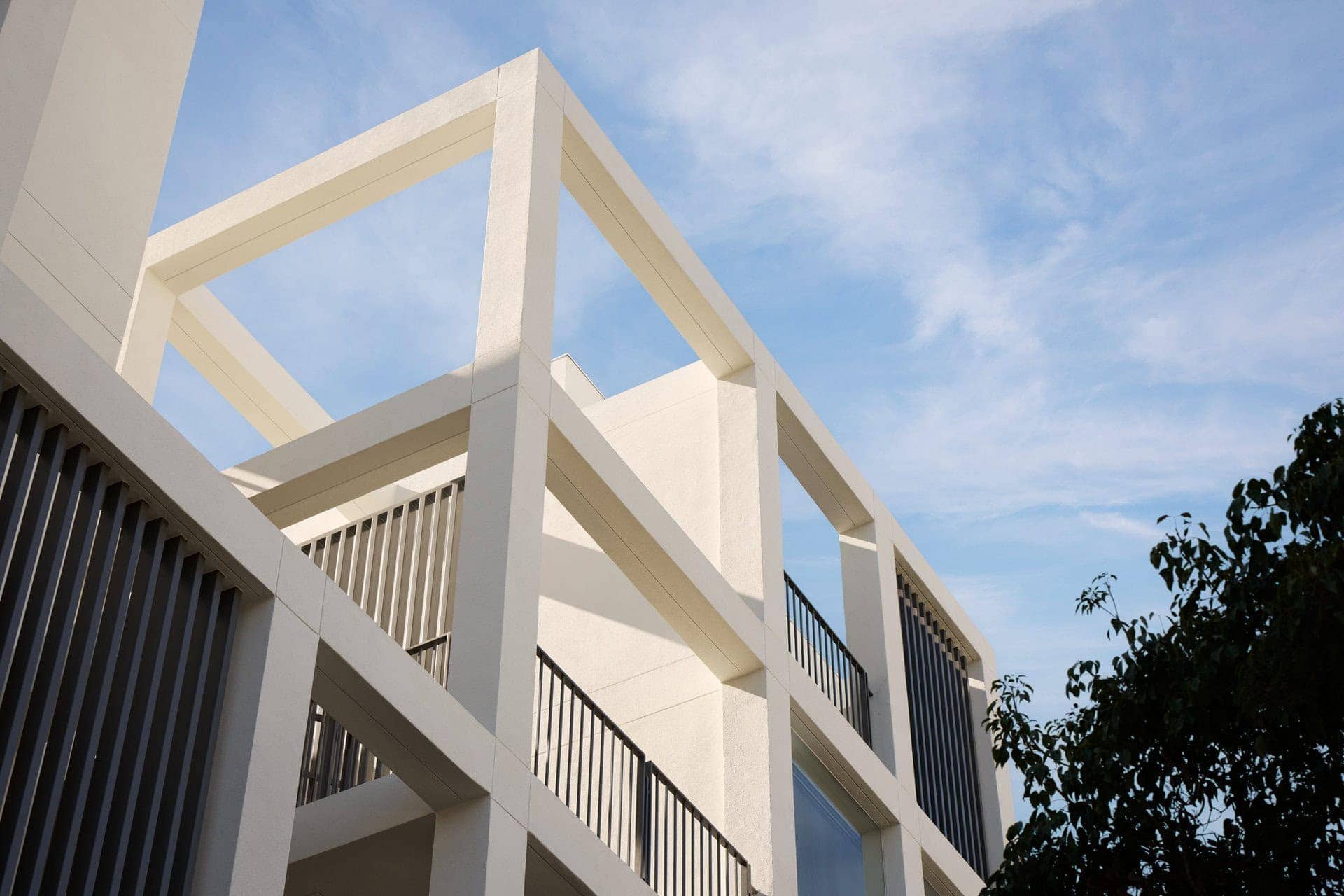 Modern white concrete building with large geometric open frames and black metal balcony railings under a blue sky.