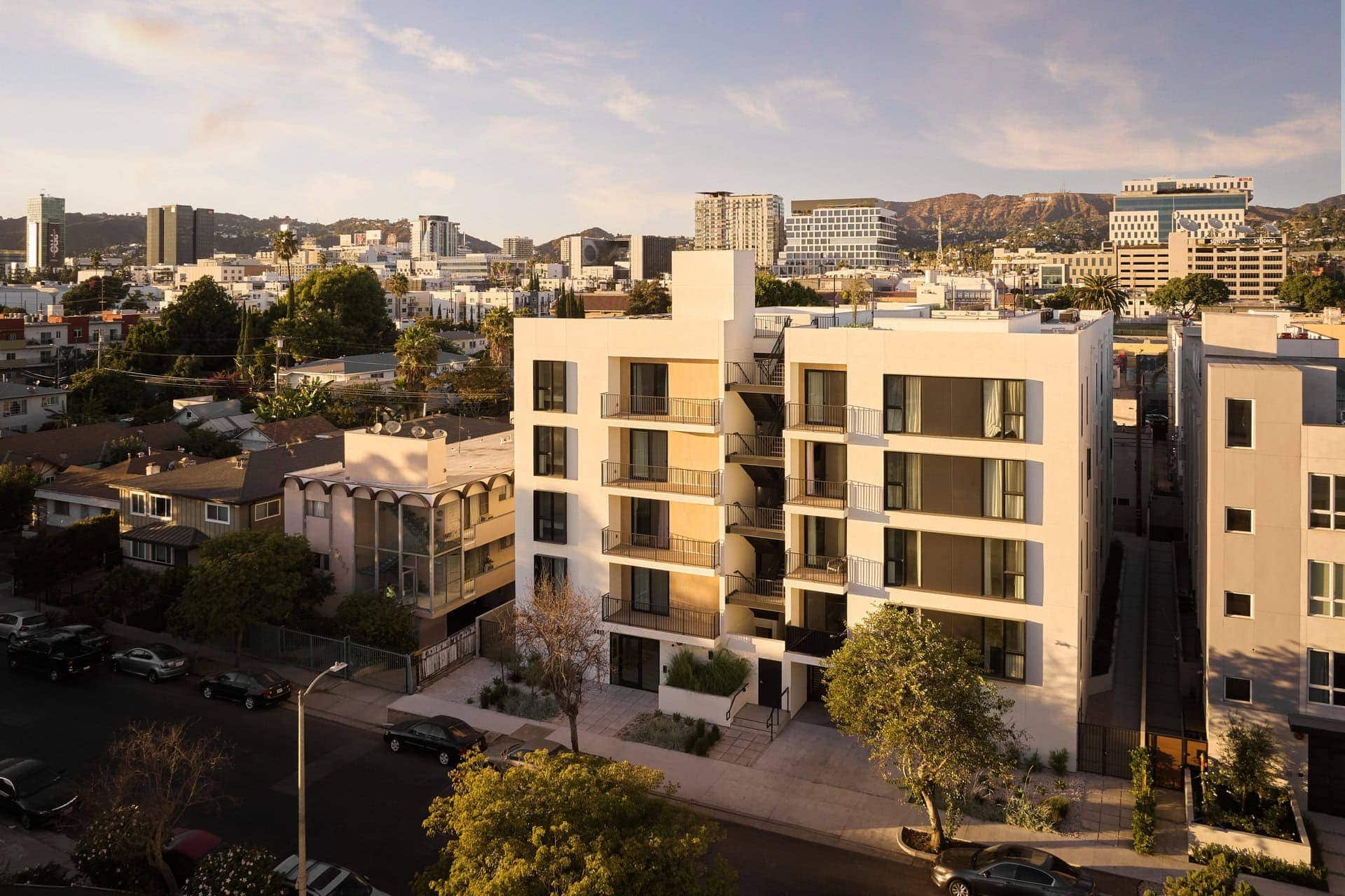 Modern white apartment building with balconies and large windows, set in an urban neighborhood at sunset.