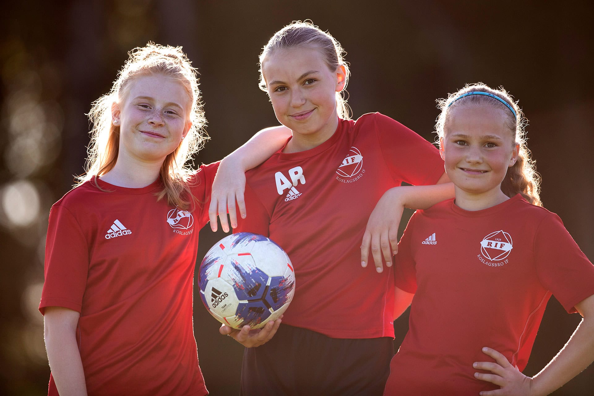 Tre flickor i röda tröjor. En av dem håller i en fotboll.