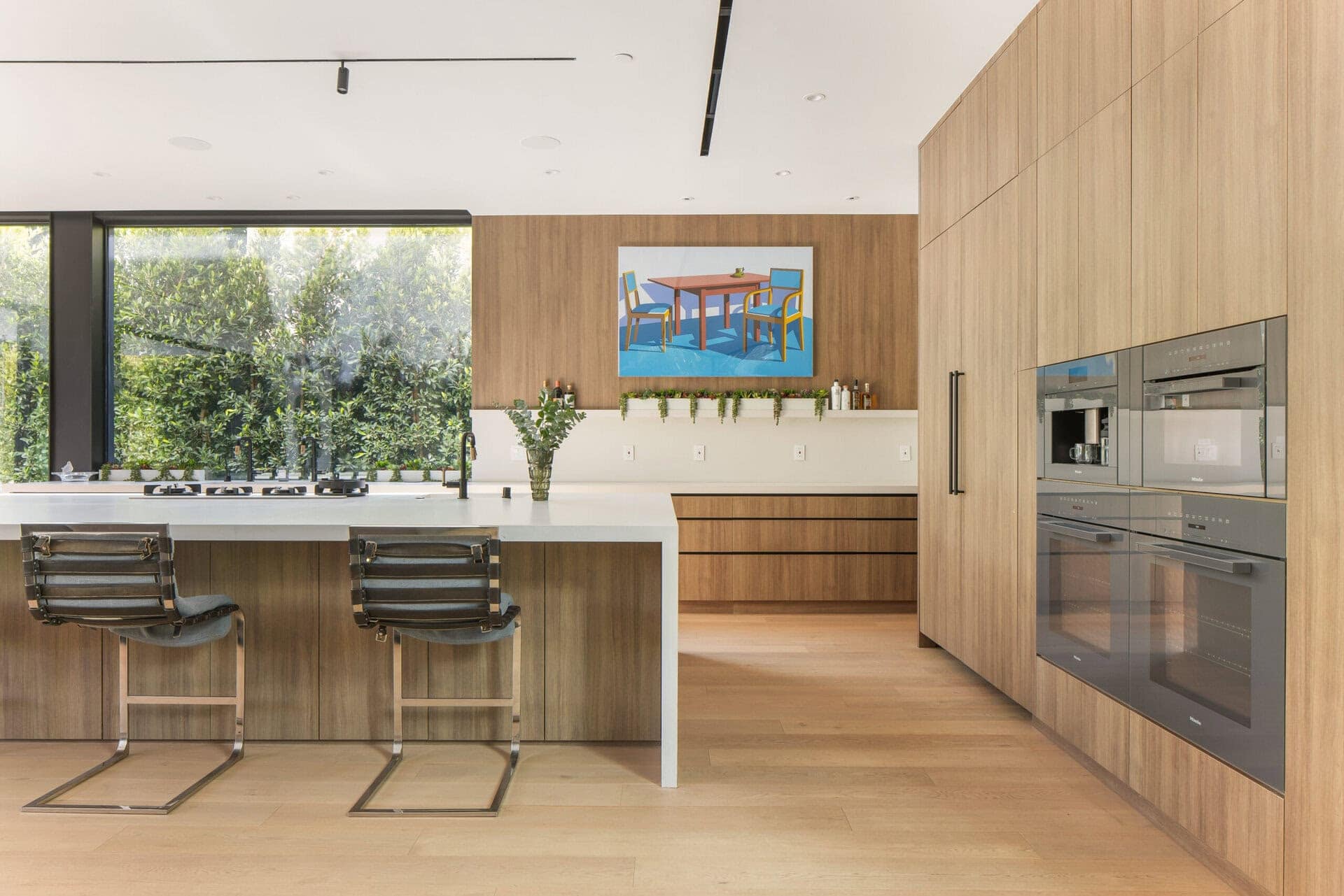 Modern kitchen with wood-paneled cabinetry and white island counter, illuminated by natural light from large windows.