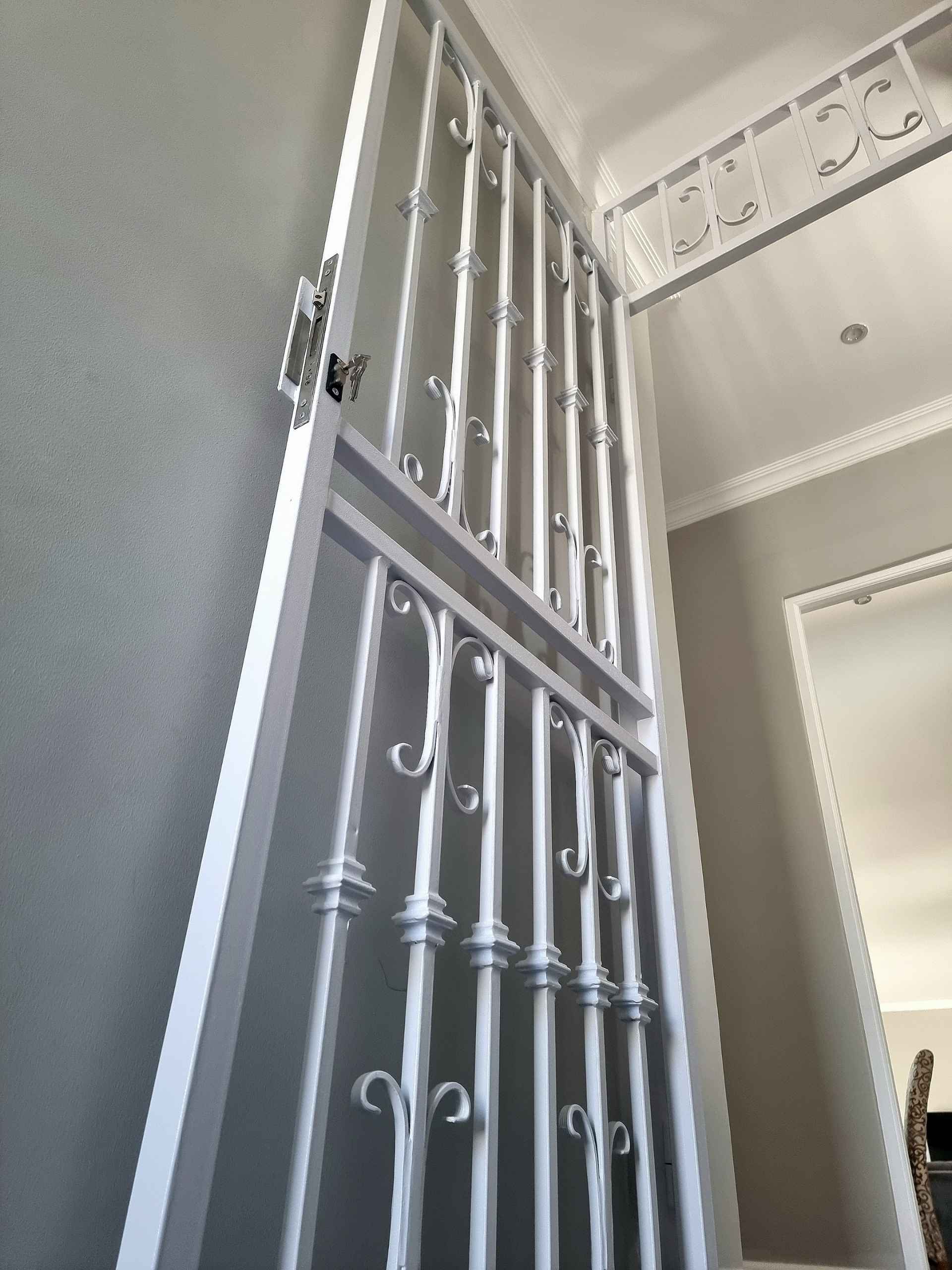 Interior white wrought-iron custom gates with decorative scroll bars acting as a full-height swing security door against a grey wall.
