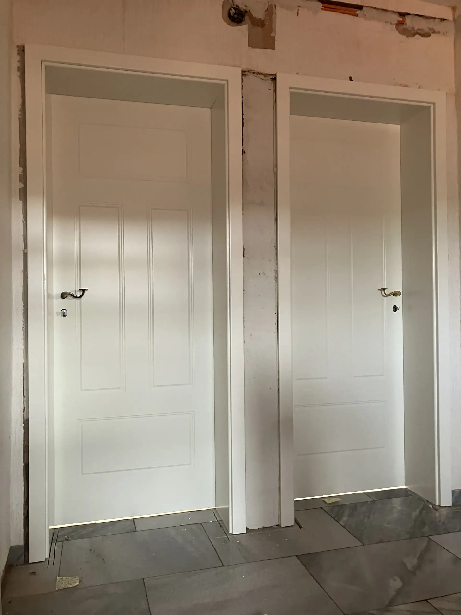 Zwei weiße Füllungstüren von Fütterer Fenster & Türen GmbH mit eleganten geschwungenen Griffen, installiert in einer Rohbauwand über dunklen Bodenfliesen.