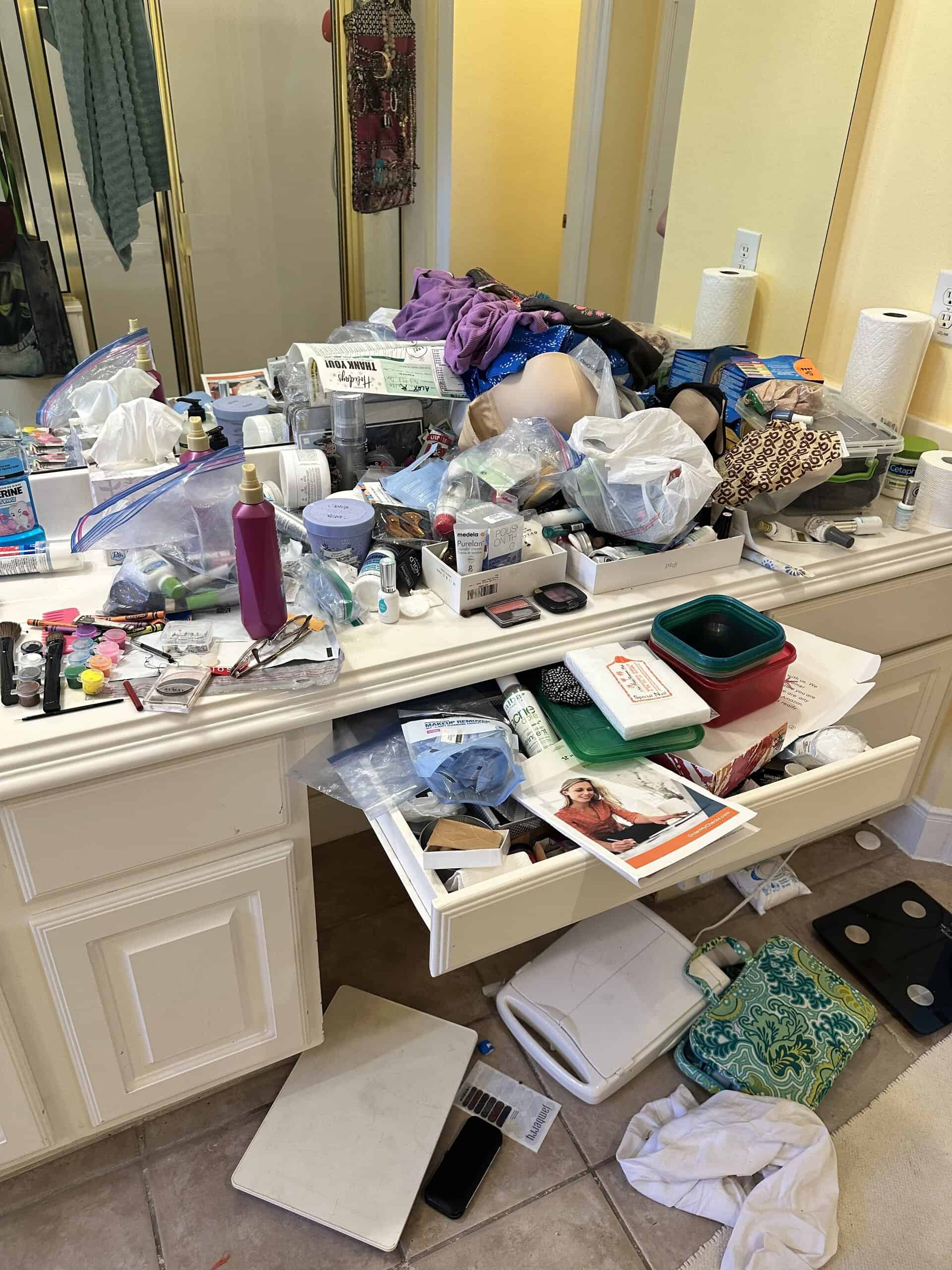 Cluttered bathroom vanity with skincare, makeup, and personal care items scattered across the surface. Includes containers, paper towels, scales, and various toiletries in a messy setting.