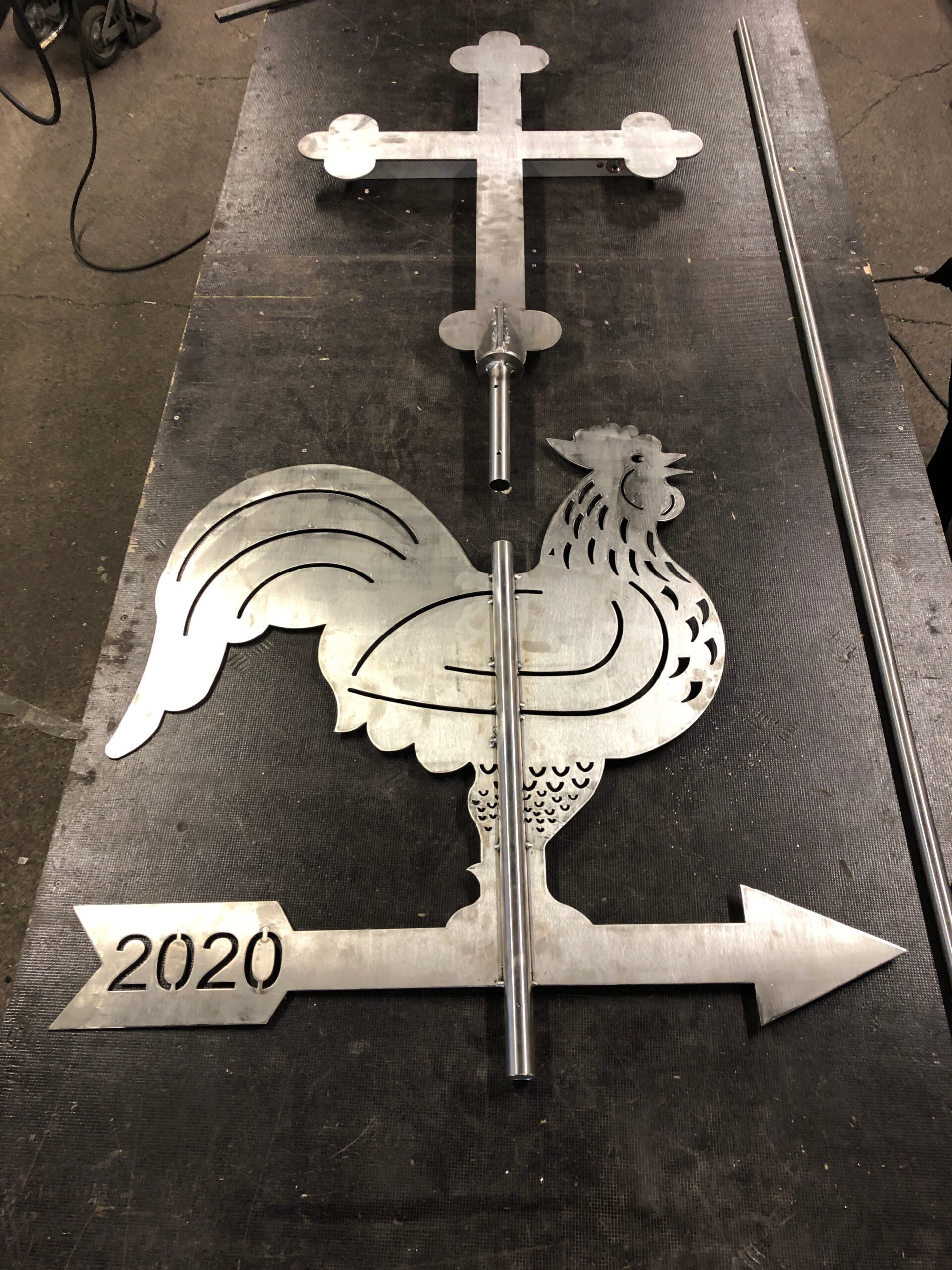 Zerlegte Wetterfahne aus Metall: Eine Hahn-Silhouette, ein Pfeil mit der Jahreszahl 2020 und ein verziertes Kreuz, bereit zur Montage. Sonderanfertigung von Stahl- und Metallbau Drost.