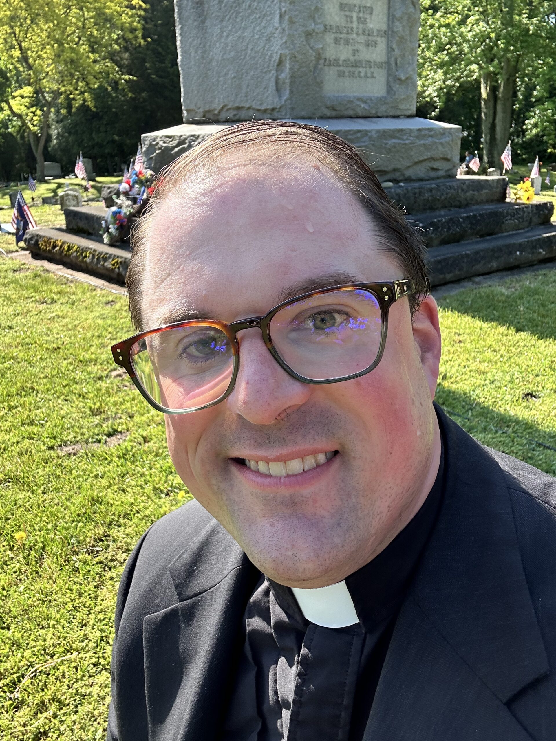 IMG_2551 - Travis Wilson A smiling man wearing glasses and a black clerical collar takes a selfie outdoors at a Memorial Day Remembrance Ceremony.