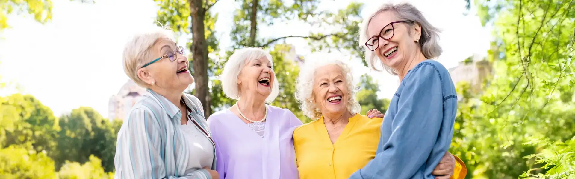 Group of four elderly women laughing together outdoors, showcasing friendship and joy in a natural setting with trees in the background.