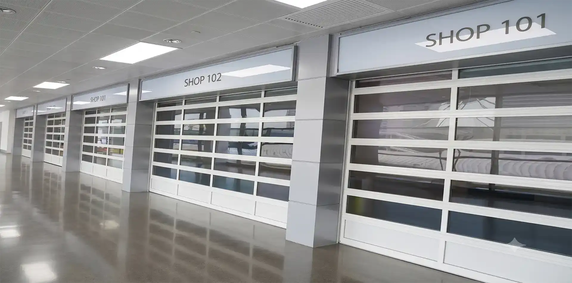 A row of white full-view glass sectional doors used as secure storefront enclosures inside a modern shopping mall, combining aesthetic appeal with robust commercial security.