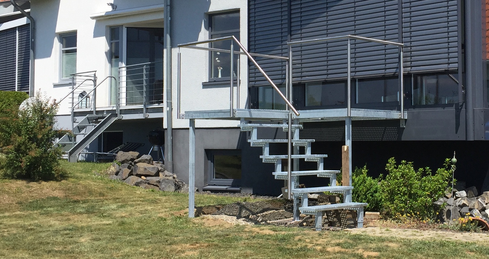 Balkon von Stahl- und Metallbau Drost: Eine robuste, verzinkte Stahltreppe mit gelochten Stufen und Edelstahlgeländer führt zu einem modernen Außenbalkon an einem Wohnhaus. Im Hintergrund ist eine weitere Stahltreppe mit Balkon sichtbar, umgeben von einem grünen Garten.