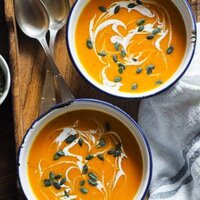 Paula's Apron Crema de Calabaza en 30 Minutos Esta Crema de Calabaza en 30 Minutos es tan reconfortante y perfecta para entrar en calor! Saludable, ligera y los toppings la llevan un pasito más allá siendo súper simples: semillas de calabaza tostadas para un contraste crujiente y un elegante pero fácil remolino de crema (o yogur) que además aporta cremosidad. En esta foto se ven 2 boles de enamel con la crema y los toppings encima de una bandeja de madera colocada en diagonal, con un trapo a la derecha y cucharas arriba a la izquierda.