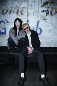 Two people sit on a bench in front of a white brick wall covered in graffiti. One person wears a gray jacket and has long dark hair, while the other wears sunglasses, a light shirt, and dark pants. Both look directly at the camera.