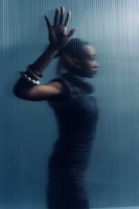 A woman stands behind a frosted, ribbed glass panel with her hand pressed against it. Her profile and features are blurred, and she wears bracelets and a dark Salma Tahir sleeveless dress. The background and lighting are cool-toned.