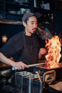 A chef at Yuzu Dining Bar in Amsterdam expertly grilling chicken over an open flame, capturing the authentic coal-fired yakitori cooking style.