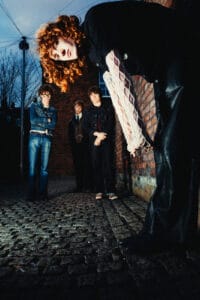 Four members of The Sukis photographed at dusk on a cobblestone street, one figure bending toward the camera with red curly hair, the others standing against a brick wall under a darkening sky — photograph by Nathan Cole