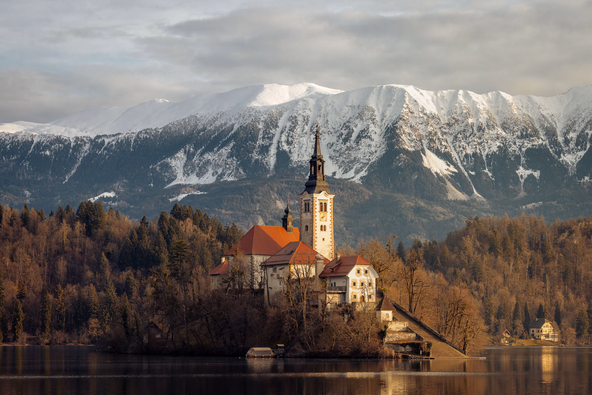 Lake Bled at sunrise with lighting dusting the snow covered mountains in background and shining on the cathedral in the center of the lake.