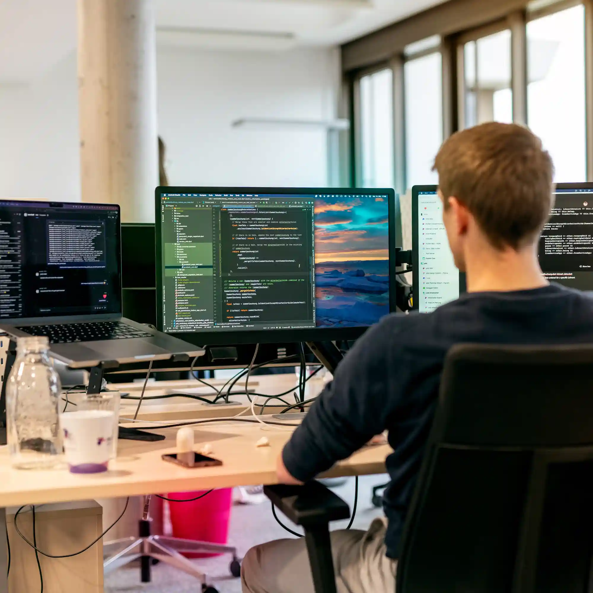 Ein Softwareentwickler sitzt an einem Schreibtisch in einem modernen Büro und arbeitet an mehreren Bildschirmen. Auf den Monitoren sind Codezeilen und eine Landschaft als Bildschirmhintergrund zu sehen. Auf dem Schreibtisch stehen eine Wasserflasche, ein Kaffeebecher und ein Smartphone.